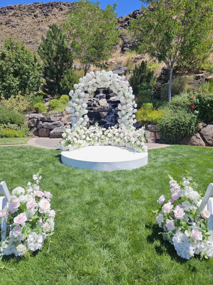 Floral archway with luxury white flowers in a garden setting with trees and waterfall.