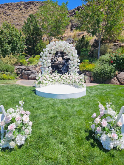 Floral archway with luxury white flowers in a garden setting with trees and waterfall.