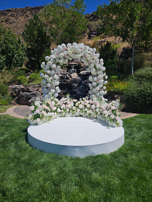 Floral arch with white flowers on a circular platform in a garden setting with trees.