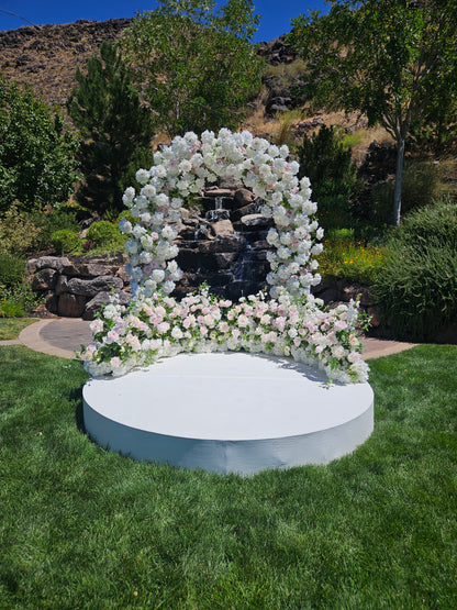 White flower wedding arch with floral arrangements on a grassy area with trees and waterfall in the background